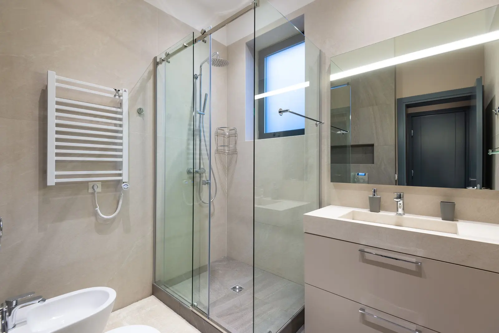Ducha con mampara de cristal corredera sobre plato gris con desagüe central, alicatado beige en gran formato, ventana pequeña de cristal translúcido, toallero blanco en la pared y lavabo con mueble a la derecha