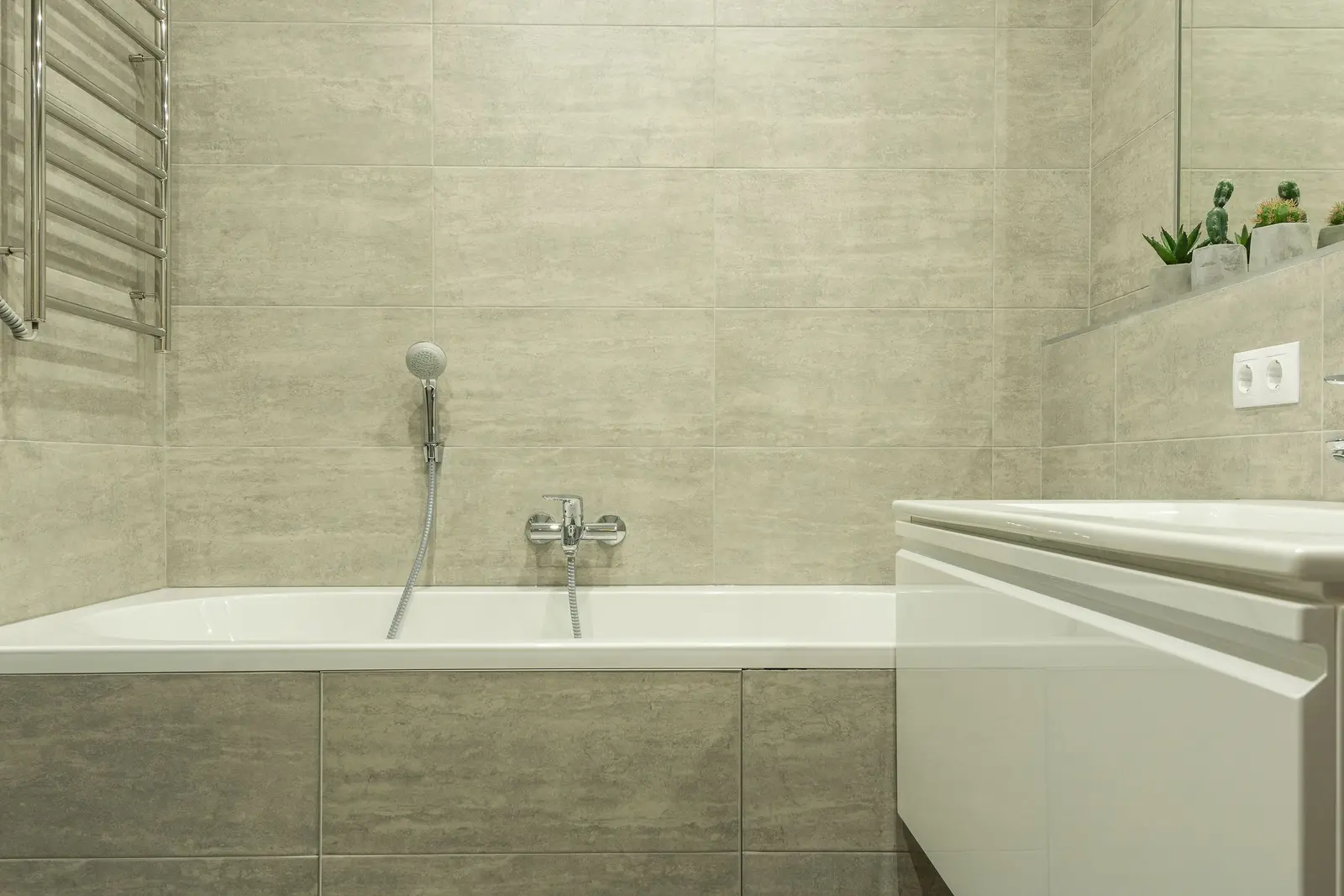 Bañera blanca empotrada con faldón alicatado en porcelánico gris claro, grifo monomando cromado y teleducha en la pared, radiador toallero a la izquierda y lavabo blanco en primer plano a la derecha