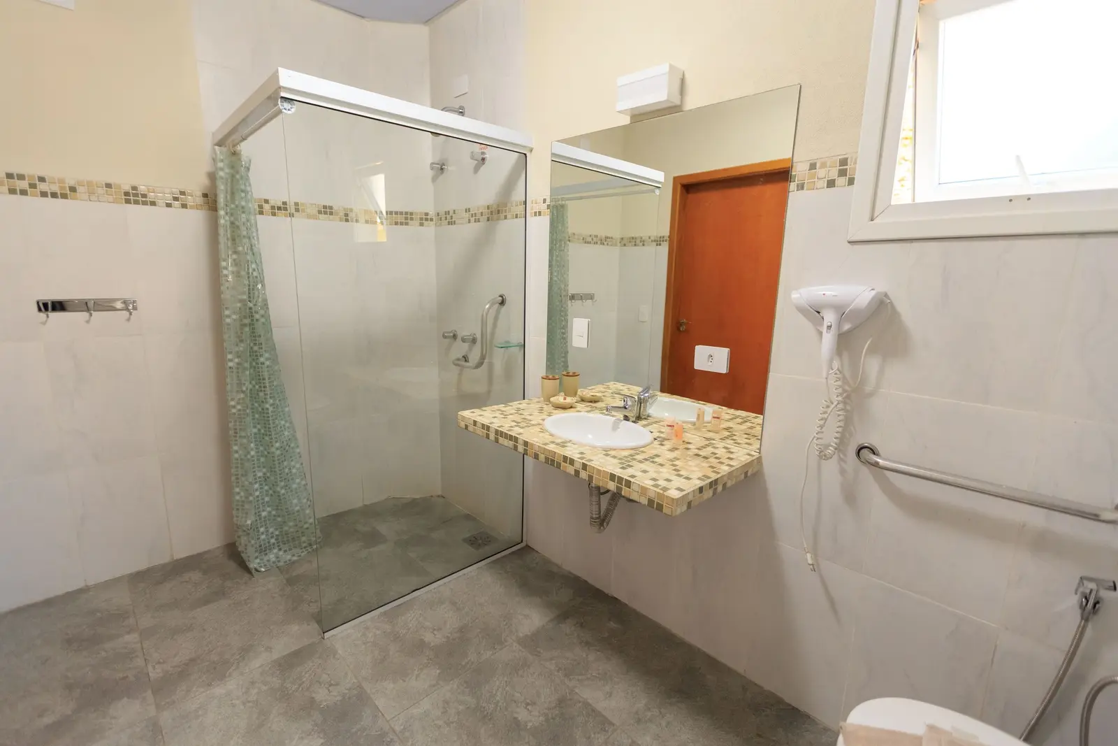 Baño amplio con ducha a nivel del suelo y mampara de cristal, asidero metálico en la pared interior de la ducha, cortina verde recogida, lavabo sobre encimera de mosaico y barra de apoyo horizontal junto al inodoro