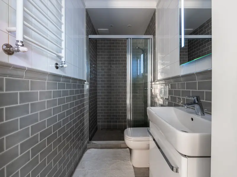 Baño estrecho con azulejo tipo metro gris en la parte baja de las paredes, ducha con mampara corredera al fondo, inodoro, lavabo con mueble blanco y toallero de agua en la pared
