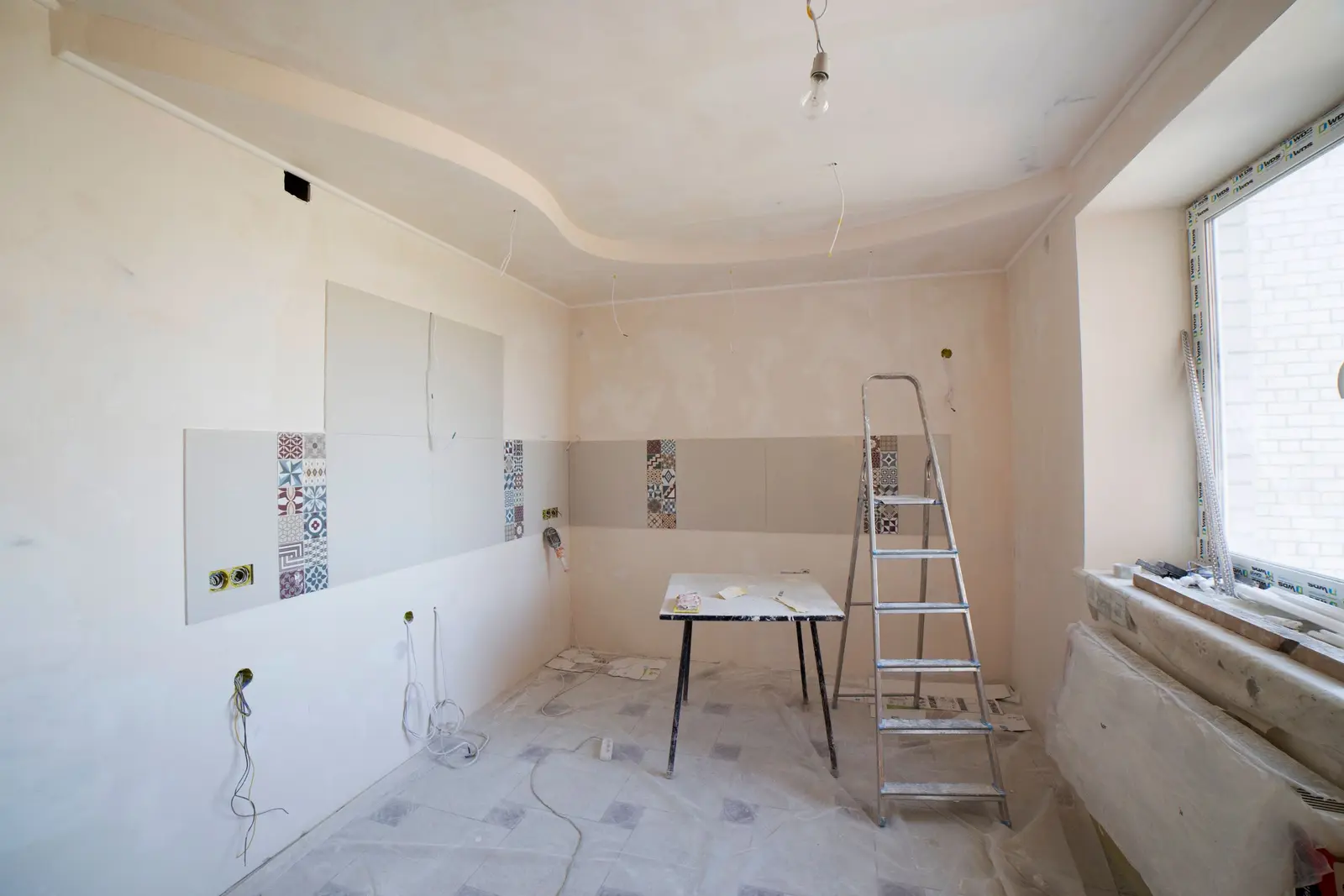 Cocina en obra sin muebles, con paredes enlucidas, cajas de enchufe y tubos de cableado a la vista, una escalera de aluminio y el suelo cubierto con plástico protector