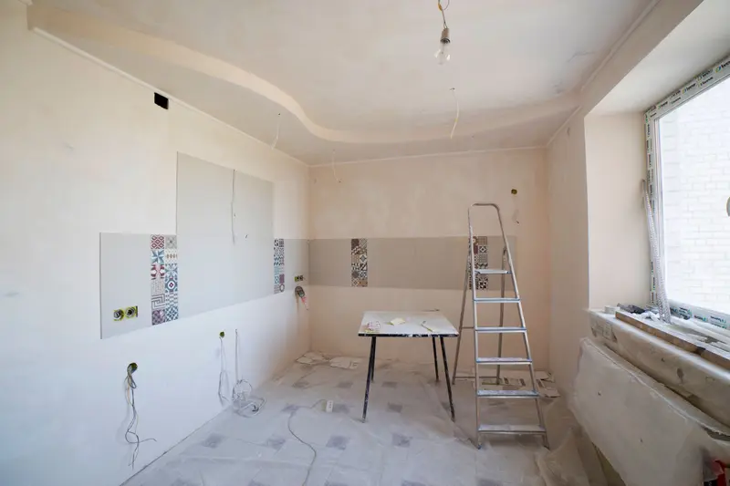 Cocina en obra sin muebles, con paredes enlucidas, cajas de enchufe y tubos de cableado a la vista, una escalera de aluminio y el suelo cubierto con plástico protector