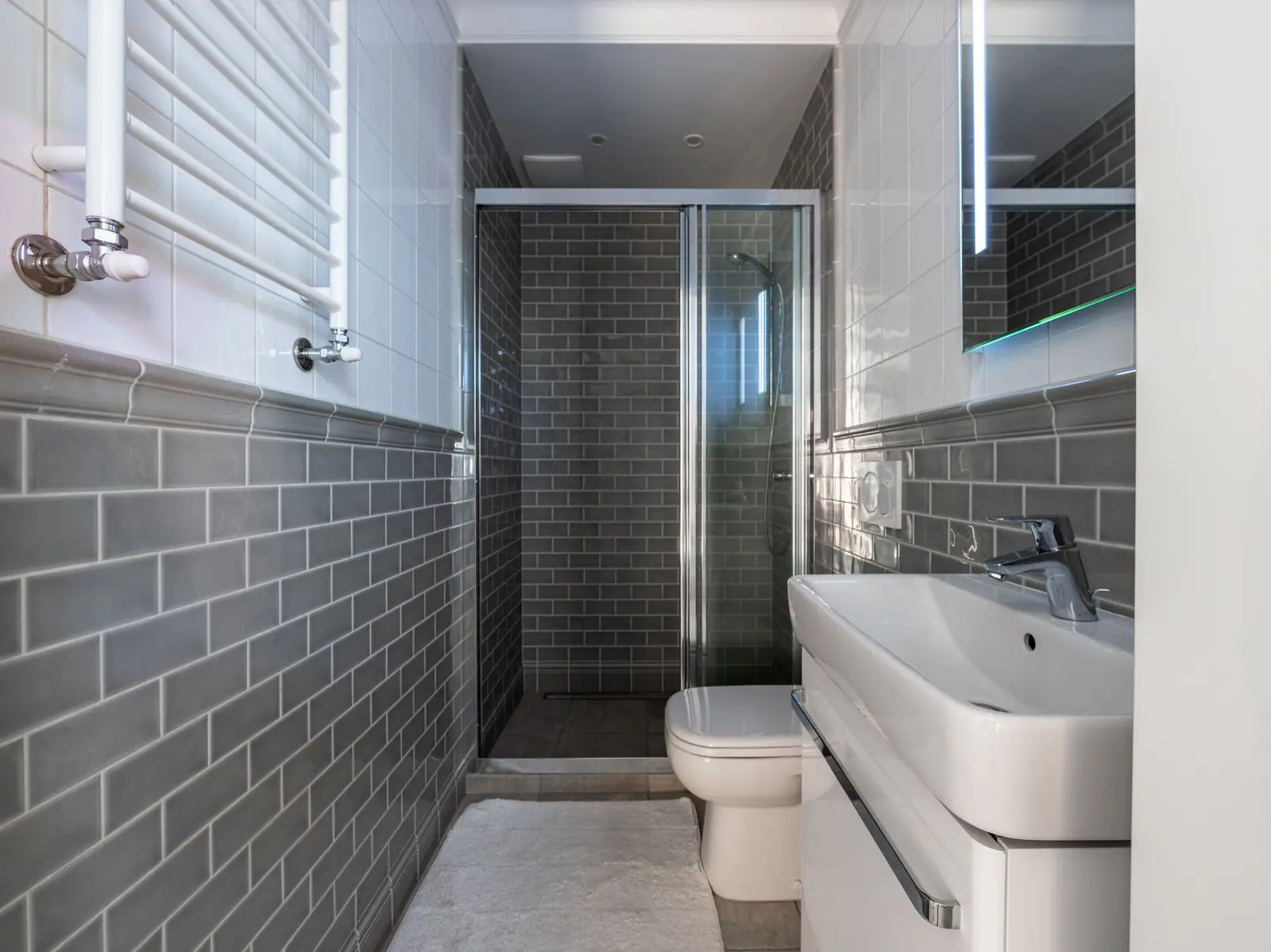 Baño alargado con alicatado gris tipo metro, mampara de cristal corredera, inodoro suspendido, lavabo con mueble blanco y toallero de pared