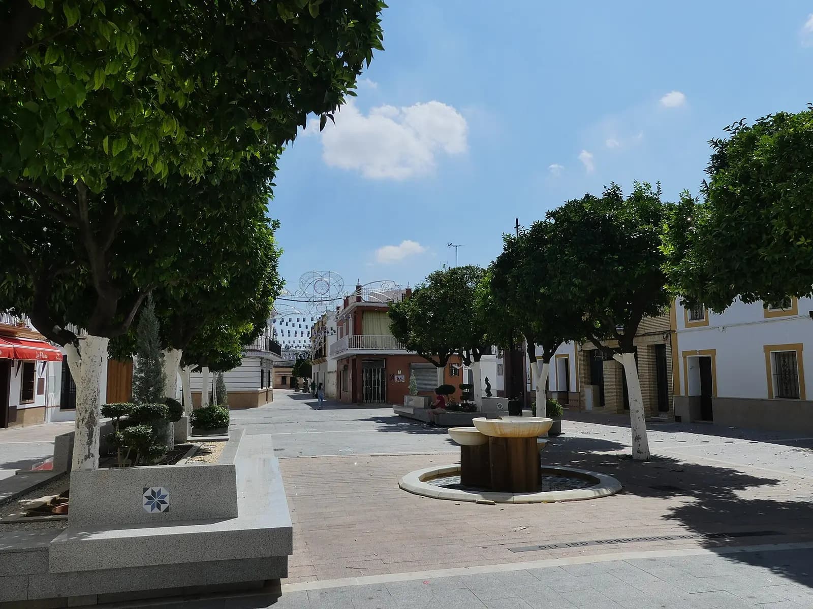 Plaza de España de La Rinconada con naranjos de tronco encalado, bancos de piedra y una fuente redonda entre casas blancas
