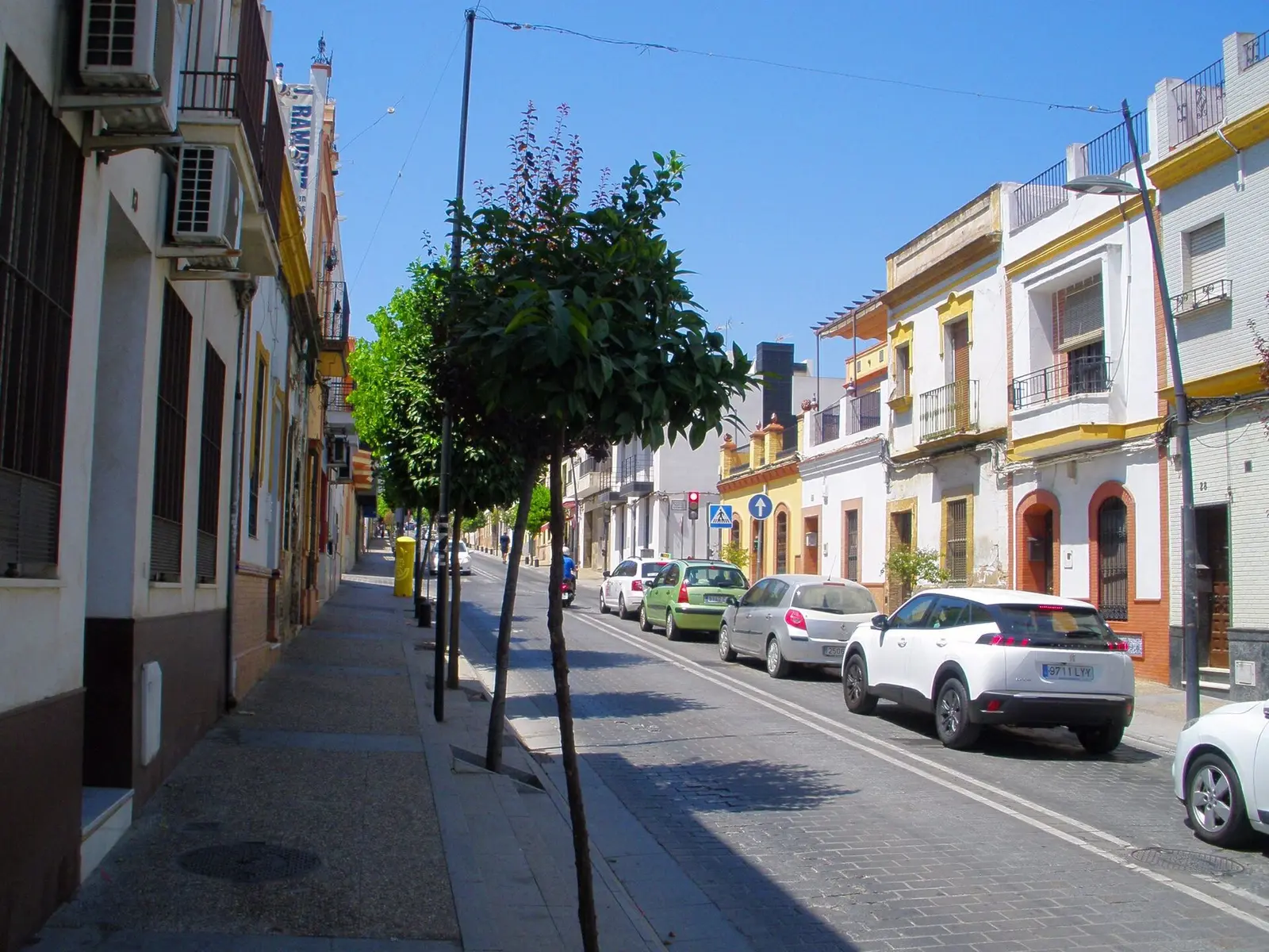 Calle de San Juan de Aznalfarache con casas de dos plantas de fachadas blancas y amarillas, rejas, aparatos de aire acondicionado en fachada, naranjos jóvenes y coches aparcados