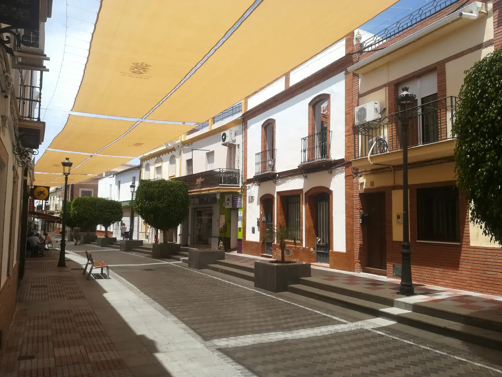 Calle Navarro Caro de Tomares, peatonal, con toldos de lona sobre la calle, casas de dos plantas encaladas con zócalo de ladrillo y naranjos en maceteros