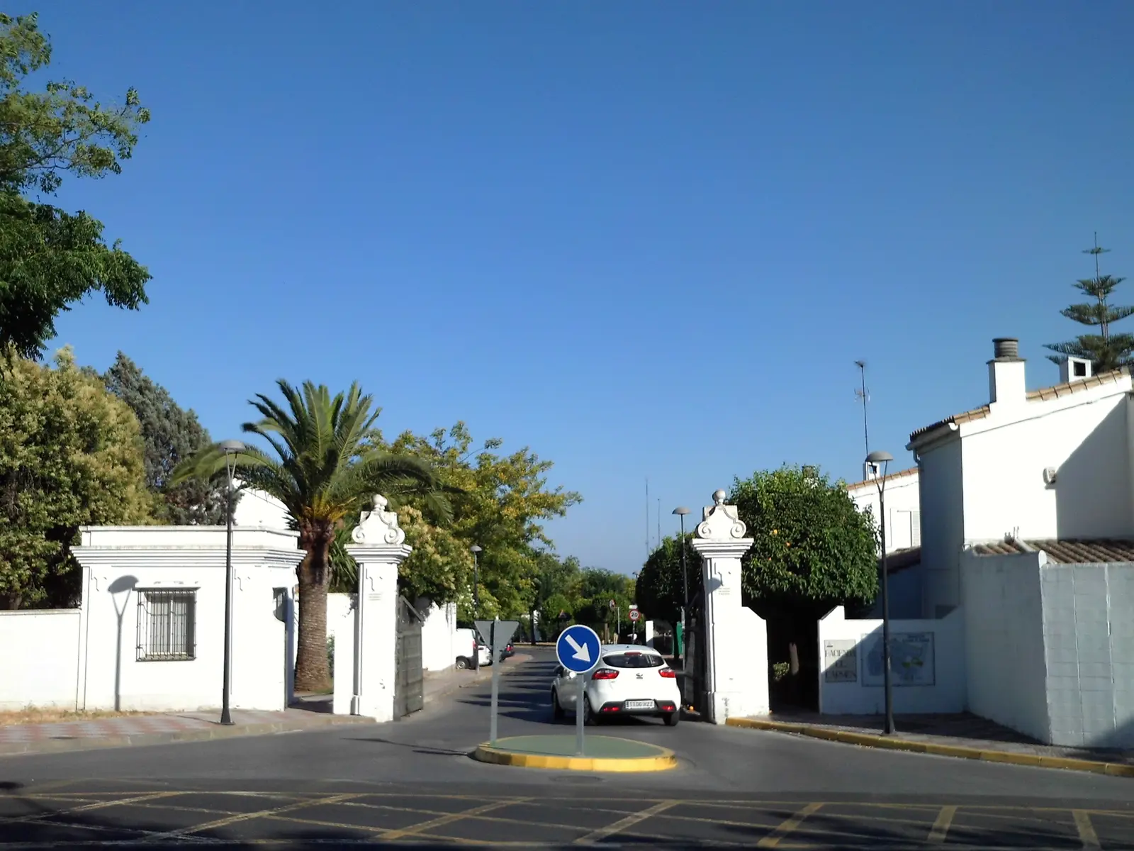 Entrada al Residencial Hacienda El Carmen en Tomares: pilares blancos con remates barrocos, una palmera, casas blancas con tejado de teja y un coche entrando